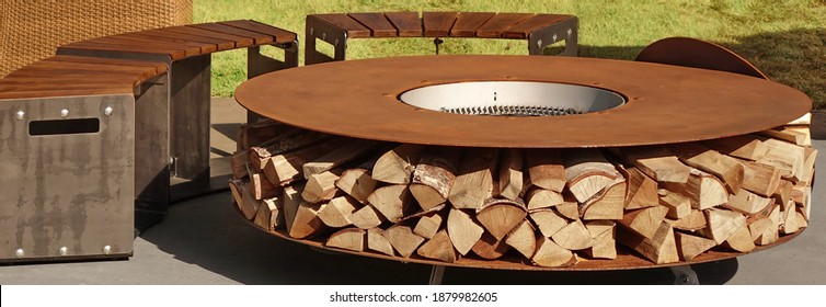 Round Patio Iron Fire Pit Table For Outdoor Leisure Party. Steel Rounded Fire Pit With Grill Top On Backyard Party Place. Grill Appliance And Fireplace On The Back Yard Lawn.