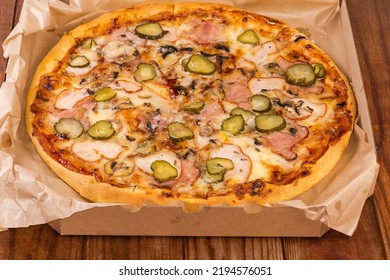 Round Pizza With Different Ham, Mushrooms And Pickled Cucumbers Served In Open Cardboard Pizza Box On The Old Rustic Table
