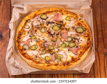 Round Pizza With Different Ham, Mushrooms And Pickled Cucumbers Served In Open Cardboard Pizza Box On The Old Rustic Table
