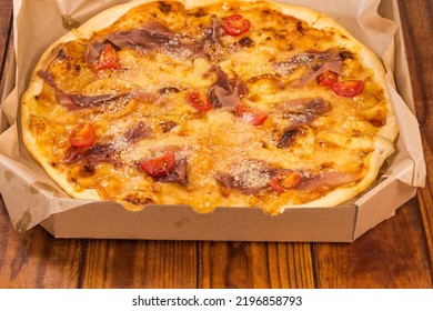 Round Pizza With Ham And Cherry Tomatoes Served In Open Cardboard Pizza Box On The Old Rustic Table, Fragment Close-up
