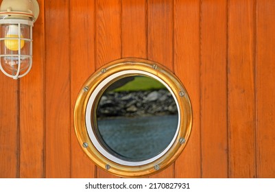 Round Port Hole On Side Of Mahogany Wall Of Boat With Shoreline Reflection.