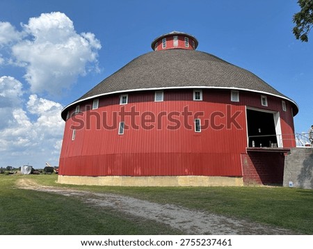 Round red barn with a dark shingled roof and cupola, set on grassy farmland under a bright blue sky. Unique rural architecture with classic Americana charm.