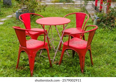 A round red table surrounded by four red chairs on the grass in a garden setting.