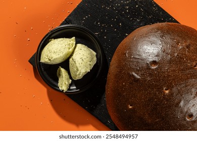 Round rye bread paired with herb butter, displayed on a black cutting board against an orange background. Ideal for bakery, food photography, or culinary-themed visuals - Powered by Shutterstock