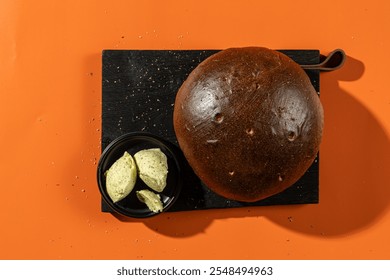 Round rye bread paired with herb butter, displayed on a black cutting board against an orange background. Ideal for bakery, food photography, or culinary-themed visuals - Powered by Shutterstock