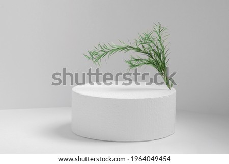 Round shape of white cylinder made of polystyrene with green twig, on light monochrome background. Empty 3d podium for presentation of cosmetic products.