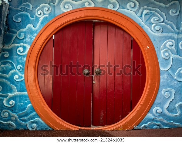 Round shaped door with orange frame and red\
wooden planks, surrounded by blue cloud sculpture. Traditional\
Chinese lion head door\
knocker.