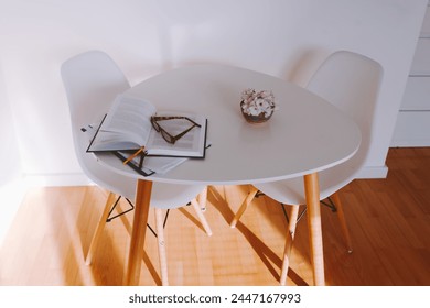 round small table on which lies an open book, reading glasses, vase flowers, chairs, near white wall, sunlight, shadows from blinds, minimalist interior, high viewing angle