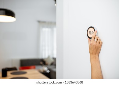 Round Smart Thermostat With Touch Screen Installed On The Wall With A Hand Trying To Reach It. Smart Home Heating Regulation Concept