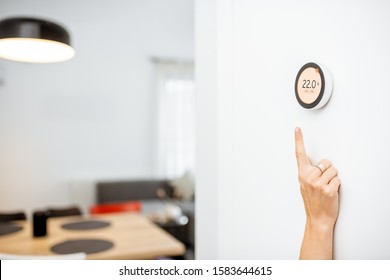 Round Smart Thermostat With Touch Screen Installed On The Wall With A Hand Trying To Reach It. Smart Home Heating Regulation Concept