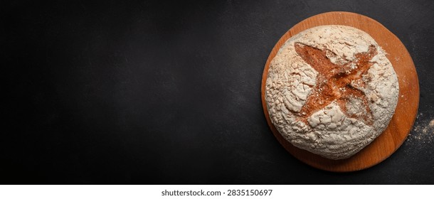 Round sourdough bread with a cross pattern sits on a wooden board, showcasing its crusty exterior and flour dust against a dark background