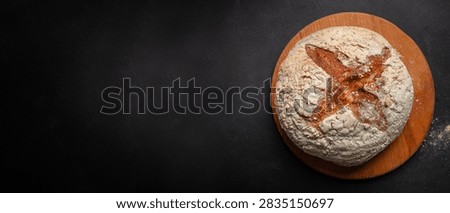 Round sourdough bread with a cross pattern sits on a wooden board, showcasing its crusty exterior and flour dust against a dark background