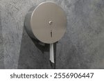 A round stainless steel toilet paper dispenser is securely mounted on a textured gray wall inside a public restroom. The dispenser has a small opening for easy paper access.