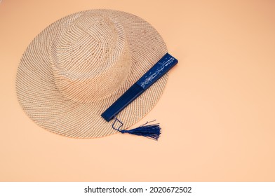 A Round Straw Hat And A Blue Hand Fan Isolated On A Peach Background With Free Space For Text- Concept For Hot Summer Weather