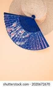 A Round Straw Hat And A Blue Hand Fan Isolated On A Peach Background With Free Space For Text- Concept For Hot Summer Weather