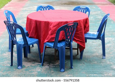 Round Table Covered With Red Tablecloth And Blue Plastic Chairs.