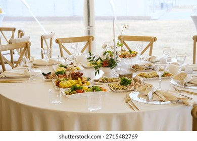 Round table with white tablecloth features plates of food, glasses, cutlery, and a floral centerpiece with various dishes arranged on it in a tent setting