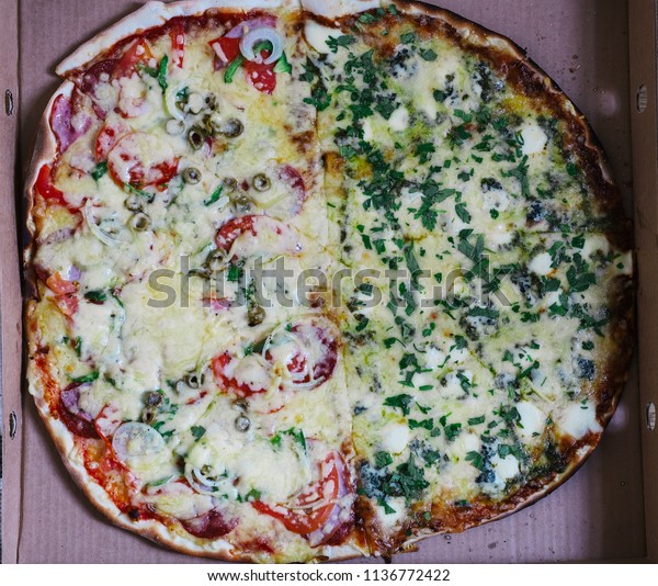 Round traditional Italian pizza on thin dough
divided into two halves - vegetarian four cheese with pesto sauce
and traditional pepperoni with olives, onion and tomatoes in a
carton box. Pizza deliver