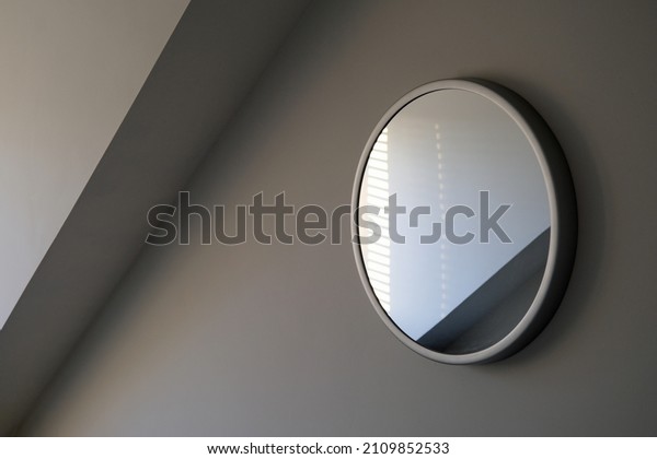 Round wall mirror reflections in a light grey room with\
angled dormer cornice. Sylish elegant and sophisticated. Simple\
enigmatic monochrome minimalist abstract interior design element.\

