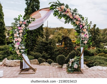 A Round Wedding Arch Decorated With Colorful Flowers And Fabric Stands In A Park, A Garden With Green Thujas In Nature. Ceremony, Photography.