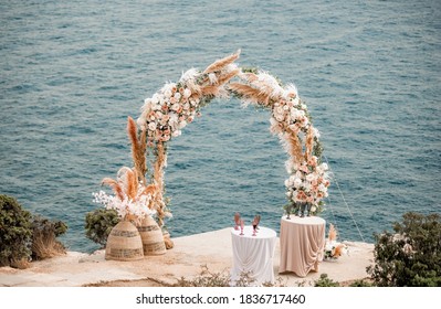 The Round Wedding Arch Is Decorated With A Variety Of Flowers For Beach Ceremony Against The Sea Landscape. Wedding Ceremony In Boho Rustic Style. Decor For Wedding Ceremony On The Sea.