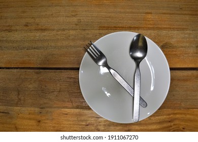 A Round White Ceramic Plate Is Placed On A Wooden Table. There Were No Food In The Plates, Only Spoon And Fork.