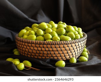 Round wicker basket filled with green olives. Around basket there are also olives. Background is black fabric. - Powered by Shutterstock