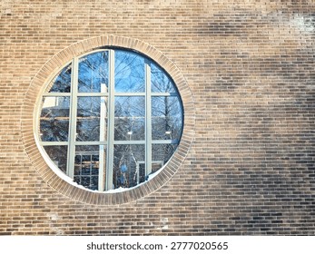 A round window surrounded by brick wall shows reflections of trees and buildings outside in an urban area during daytime.