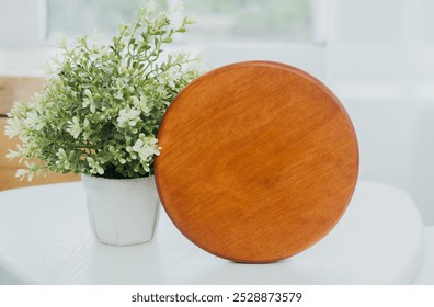 A round wooden plate rests next to a small potted plant on a white table in natural light. - Powered by Shutterstock