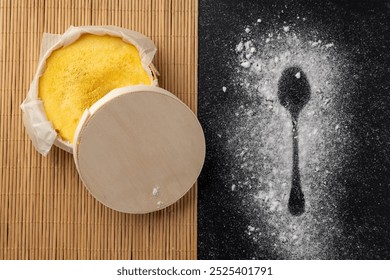 A round yellow cheese in a wooden box with a lid rests on a bamboo mat; beside it on a black surface, sugar powder sprinkled in the shape of a spoon creates intrigue. - Powered by Shutterstock