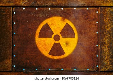 Round yellow radioactive (ionizing radiation) danger warning symbol painted on a massive rusty metal plate fixed with metallic screws to the wall and with dark rustic grungy texture background.