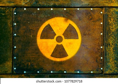 Round yellow radioactive (ionizing radiation) danger warning symbol painted on a massive rusty metal plate fixed with metallic screws to the wall and with dark rusty brown and green moss texture.