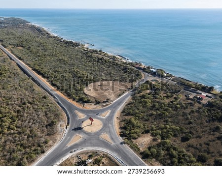 A roundabout sits at the intersection of two roads along the coast. The sea is visible at one side and trees surround the area.