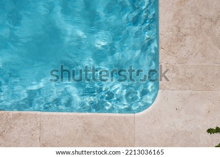 rounded corner of pool with light blue water, stone edges or light beige tiles. A small piece of plant branch sticking out at the bottom right of the picture