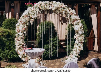 
A round-shaped wedding arch, decorated with white and pink flowers, a green leaf, and crystal pendants whistle on the arch. Crystal curtain. Wedding ceremony in the green garden. Wedding of the bride