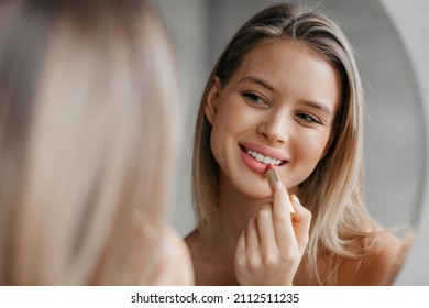 Routine procedures and decorative cosmetics for homemade visage. Happy young woman applying matte lipstick in bathroom interior, looking in mirror, closeup