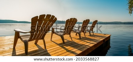 Row of Adirondack chairs -patio - deck chairs on wooden dock with sunset or sunrise -cottage life. Banner - panoramic