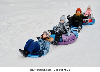 Row of adorable happy children in winter jackets sitting on slides and snow tubes and riding down hill in natural environment - Powered by Shutterstock