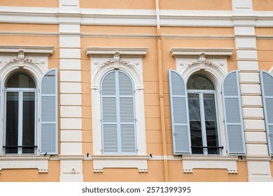 Row of arched windows with open and closed wooden shutters painted white in yellow wall, view outdoors in overcast day