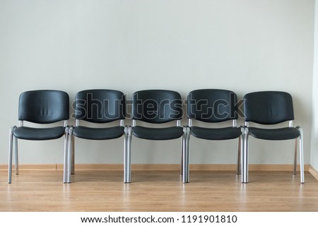 Row of black office chairs standing in corridor or conference room, empty dark seats arranged in line in boardroom, range of basic stools in front of white wall. Interview, recruitment concept
