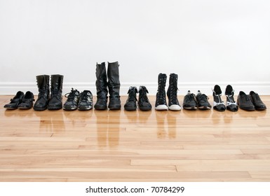 Row Of Black Shoes And Boots On A Wooden Floor, In Front Of A White Wall.