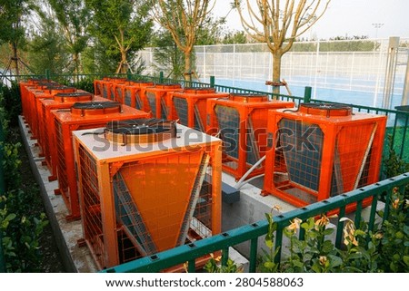 A row of bright orange air source heat pump units installed outdoors, surrounded by green fencing and trees.