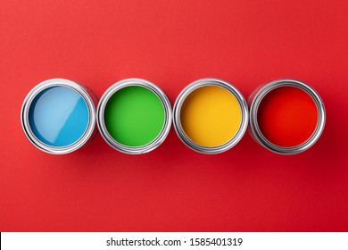 Row Of Cans Of Colorful Paint On Red Background. Top View On Green, Yellow, Red And Blue Paints.