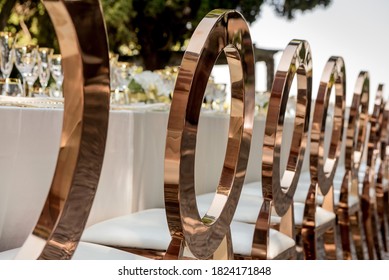 Row of chairs with shiny, brass-coloured, oval backs neatly lined in a row along the length of a long table set with elegant wine glasses and tableware