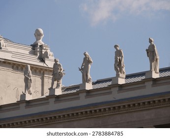 Row of classical statues standing on the rooftop of a historic building, silhouetted against a bright blue sky with soft clouds. Taken in front of the Hungarian Parliament in Budapest.