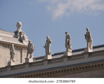 Row of classical statues standing on the rooftop of a historic building, silhouetted against a bright blue sky with soft clouds. Taken in front of the Hungarian Parliament in Budapest.