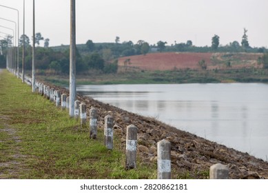 Row of concrete boundary posts along a grassy dam crest overlooking a calm reservoir and rocky slope