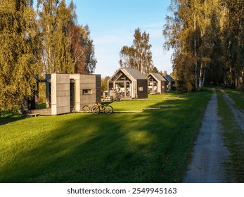 A row of contemporary cabins with large windows sits along a grassy path. Bicycles are parked outside, surrounded by tall trees and a clear blue sky. - Powered by Shutterstock