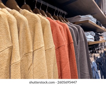 Row of cozy knit sweaters in warm earthy tones hanging on wooden hangers in a retail clothing store. Neatly organized display with folded jeans and shirts stacked on shelves above and below. - Powered by Shutterstock - Get 15% off with code: PIKWIZARD15