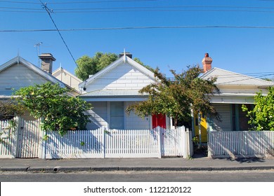 Row of detached bungalow homes in the residential suburb of St Kilda in Melbourne with a white picket fence and a blue sky background. Power cables overhead. 